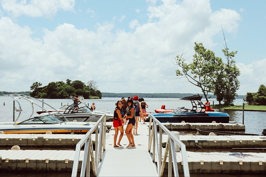 Two campers standing on a dock on a lake line up to board a boat on a sunny day.