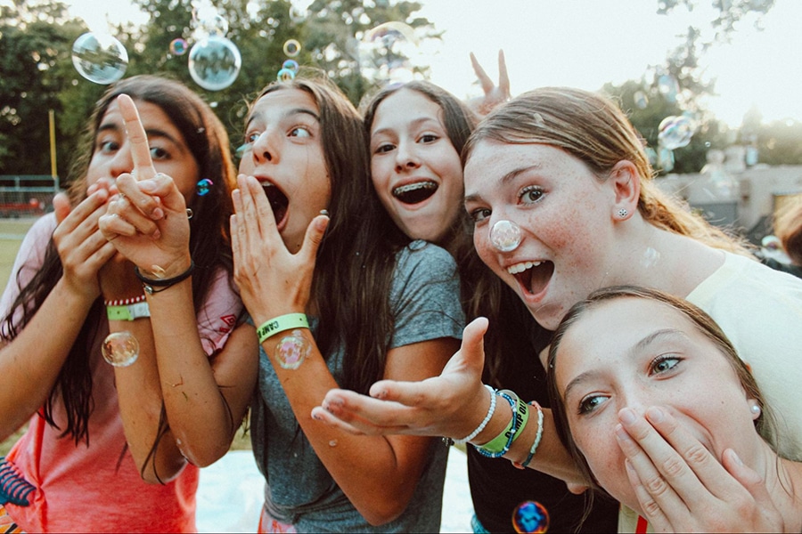 A group of five campers smile and laugh while bubbles float by on a sunny afternoon.