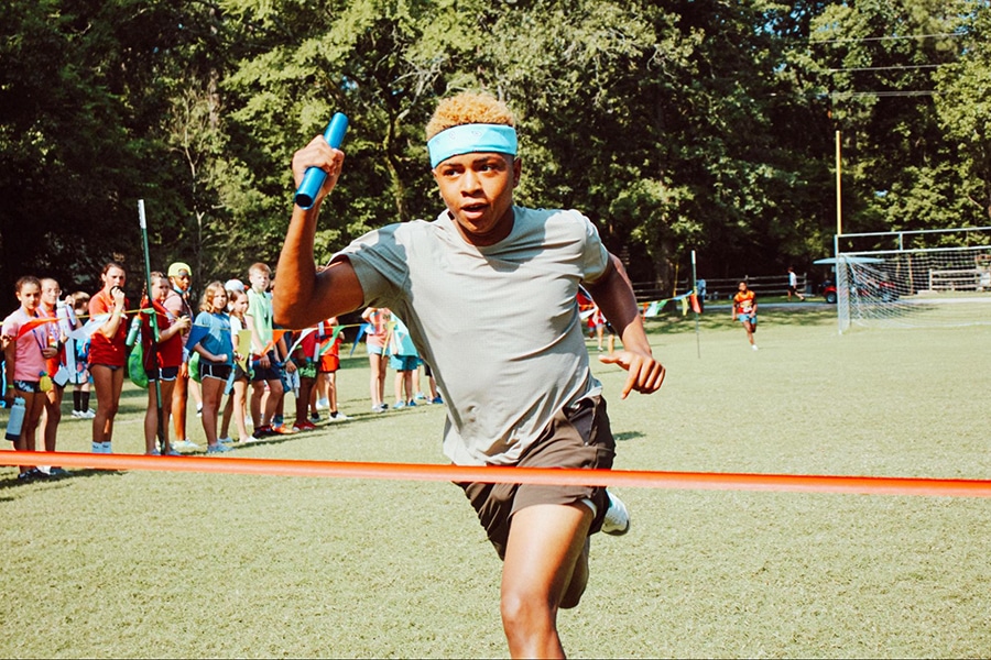 A camper wearing a sweatband and holding a baton runs across the finish line during a race.