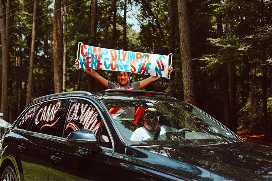 A smiling father drives by a summer camp entrance while his son stands in the open sunroof holding a Camp Olympia banner.