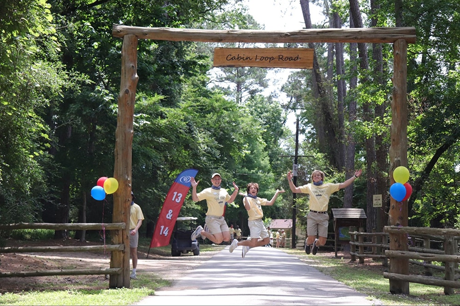 Four smiling summer camp counselors jump in the air at the Camp Olympia Cabin Loop Road entrance gate.