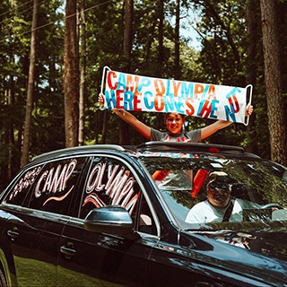 A smiling father drives by a summer camp entrance while his son stands in the open sunroof holding a Camp Olympia banner.