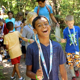 A group of children and instructors interact on a trail in the woods during a nature hike.