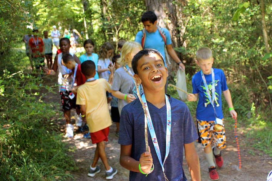 A group of children and instructors interact on a trail in the woods during a nature hike.