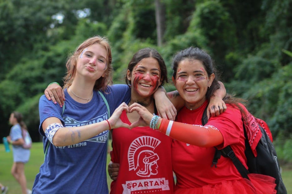 Three smiling girls at Camp Olympia making a heart sign with their hands.