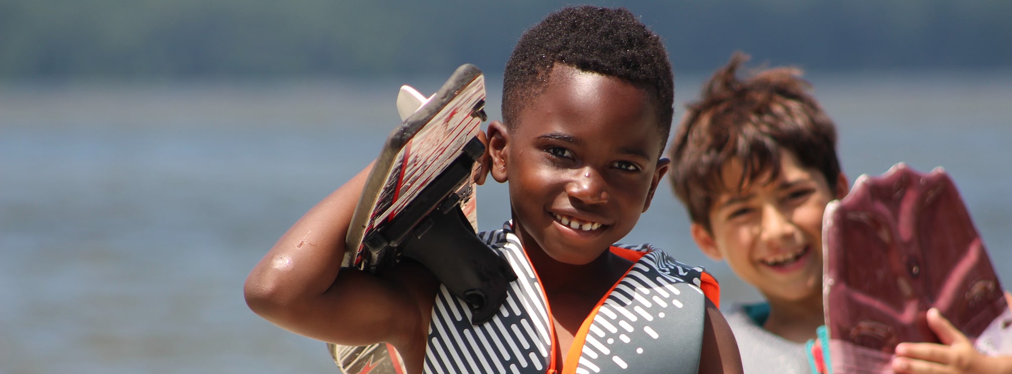 Two Camp Olympia campers carrying a waterski and wakeboard with Lake Livingston in the background.