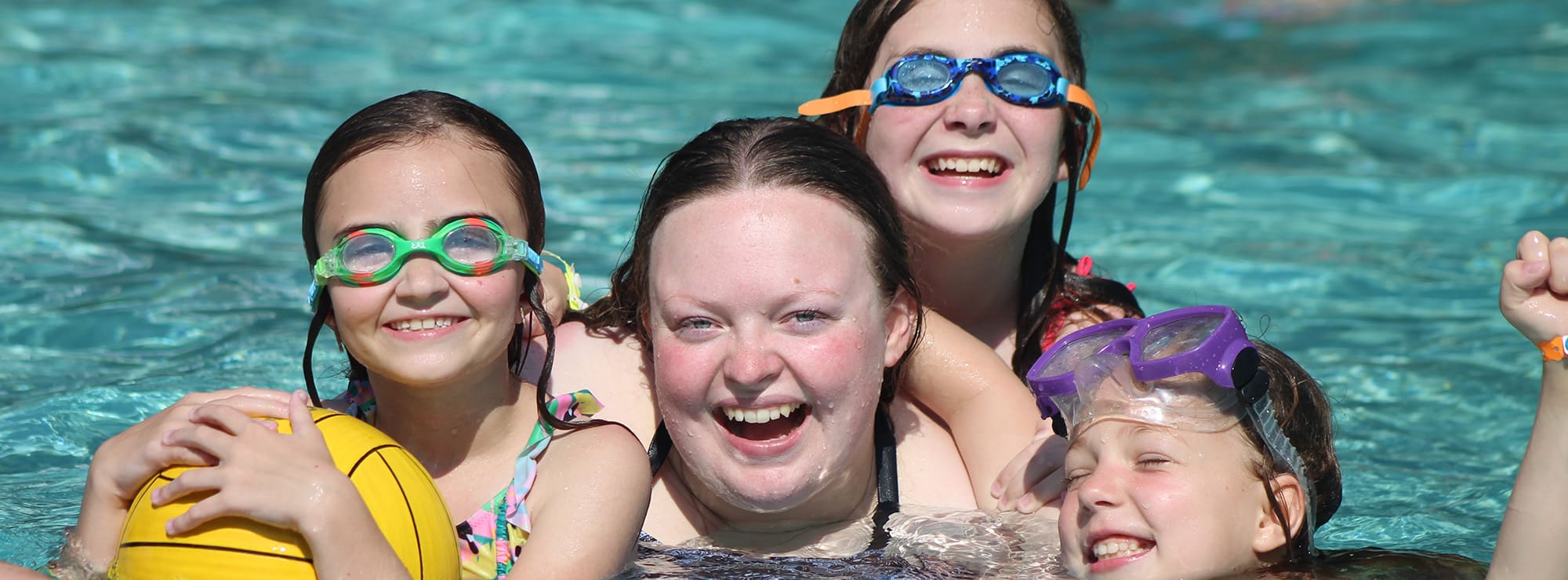 A summer camp counselor smiling in the camp pool with three girl Camp Olympia campers on a sunny day.