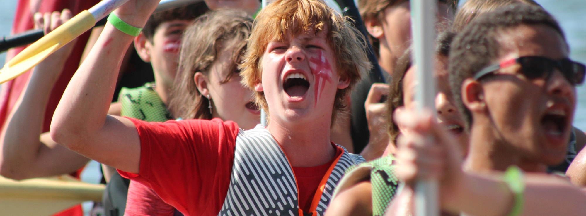 Camp Olympia camper with red face paint cheering while holding a canoe paddle.