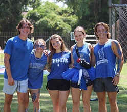 A group of five smiling camp counselors in blue Athenian wear posing together outdoors on a sunny day.