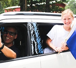 A parent driving a white SUV and a smiling young camper holding a Camp Olympia flag from the open windows.