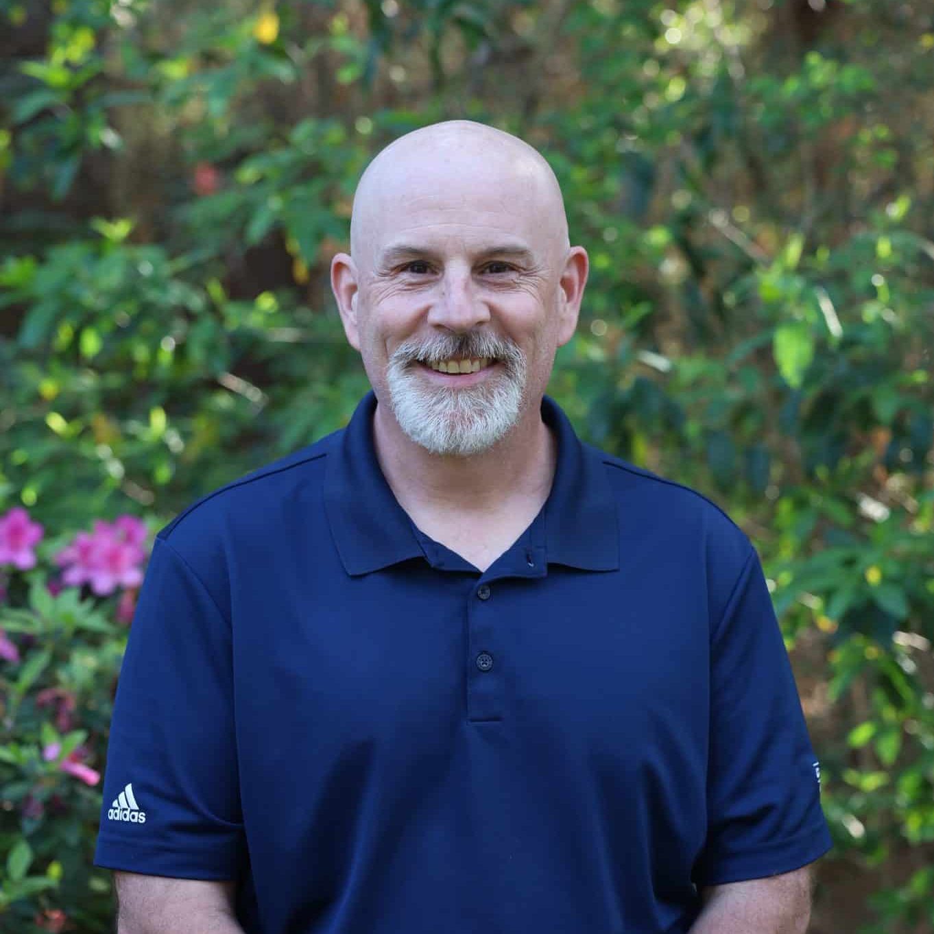 Bald man with a gray beard smiling, wearing a navy blue polo shirt, standing outdoors with greenery behind him.