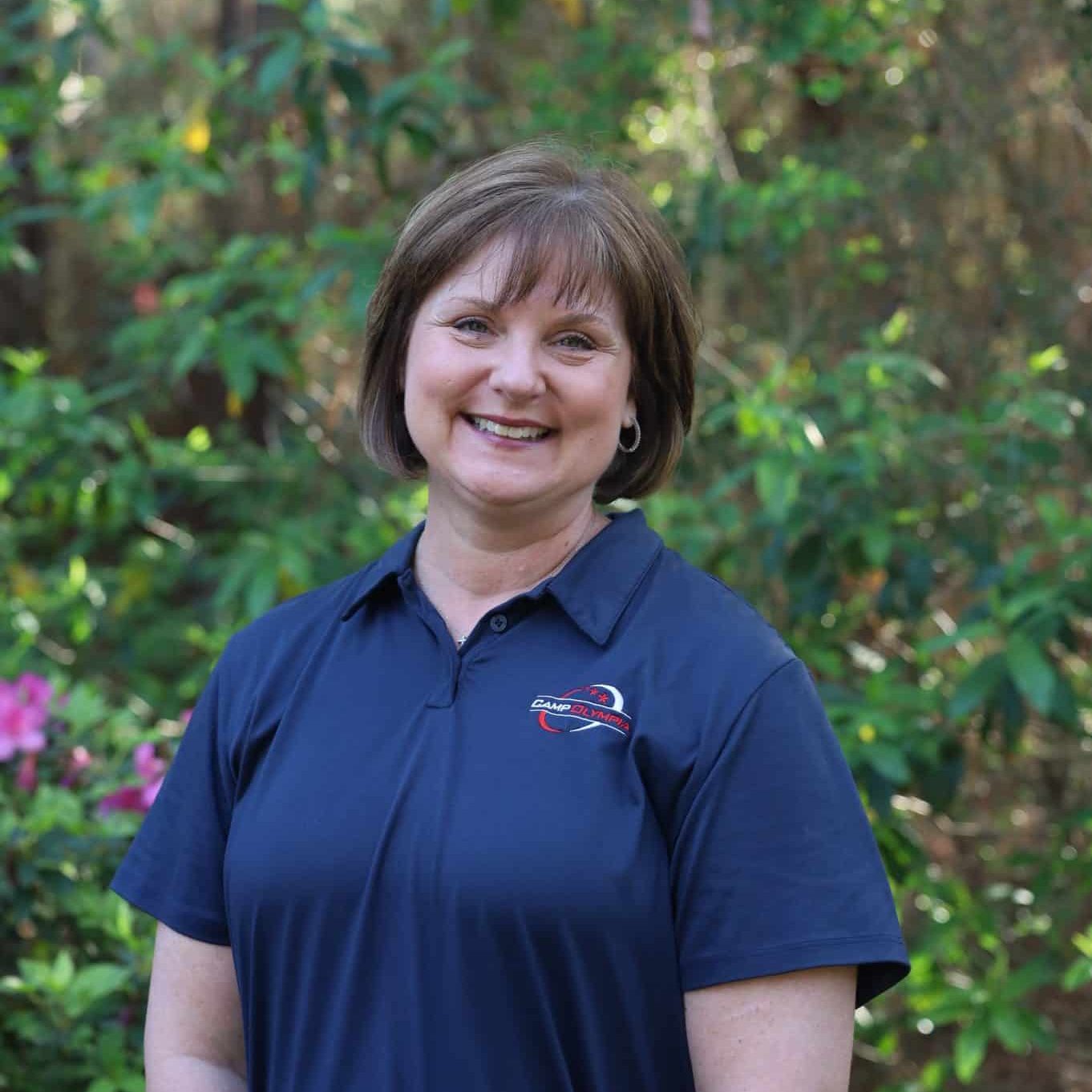 Woman smiling outdoors in a navy polo shirt with a logo, standing in front of green foliage and flowers.