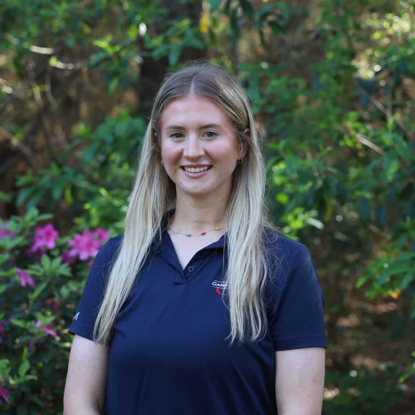 Woman with long blonde hair in a navy polo shirt smiling, standing outdoors with green plants and pink flowers behind her.