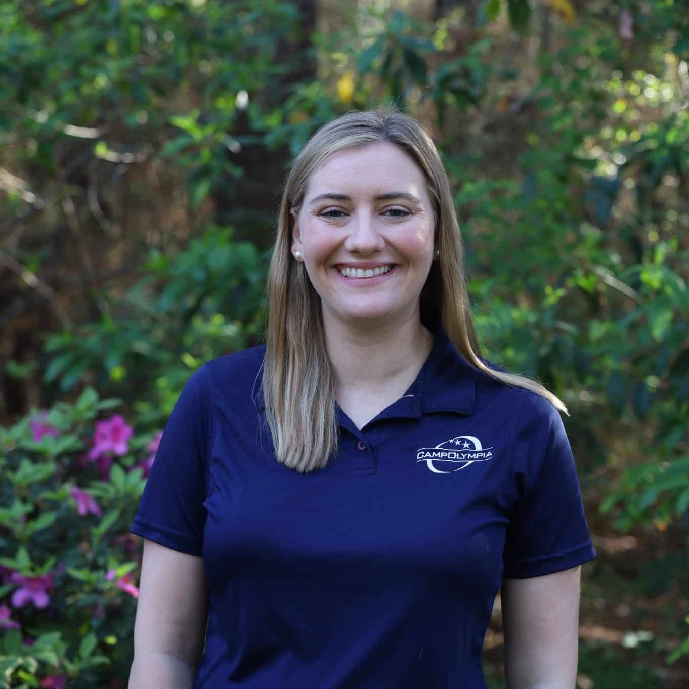 A woman smiles outdoors, wearing a navy polo shirt with a Camp Olympia logo, surrounded by greenery.