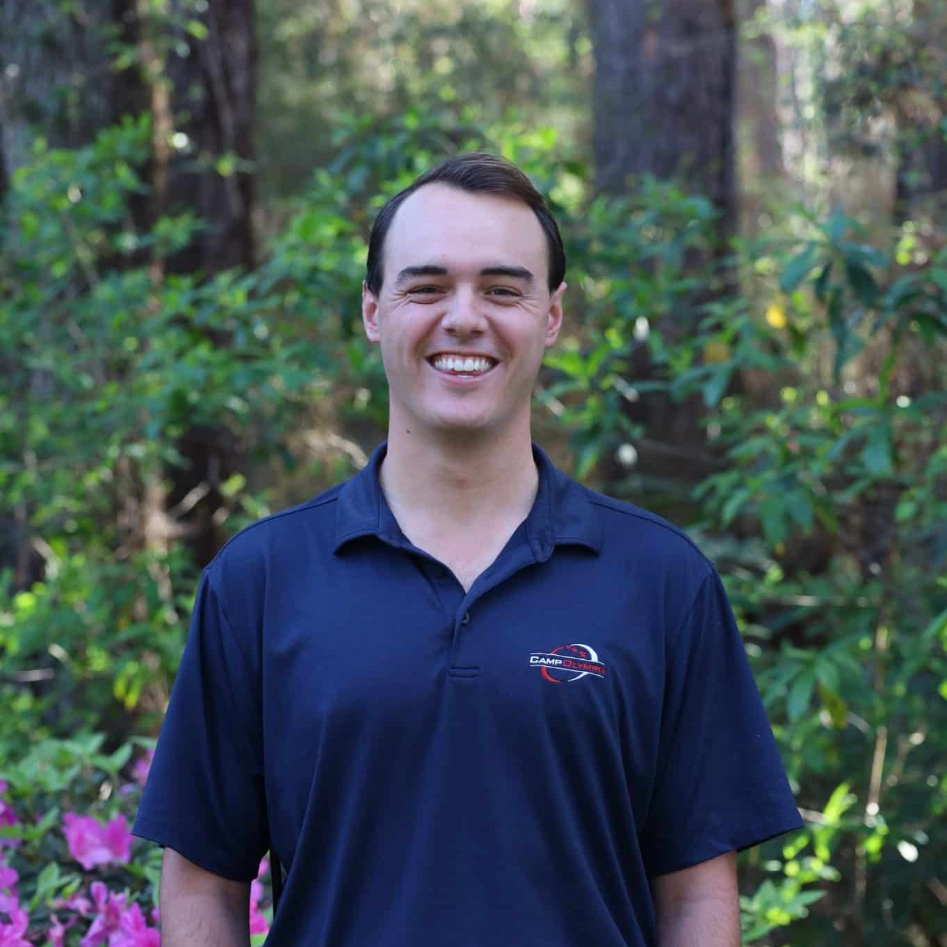 A smiling man in a navy polo shirt stands outdoors with greenery and pink flowers in the background.