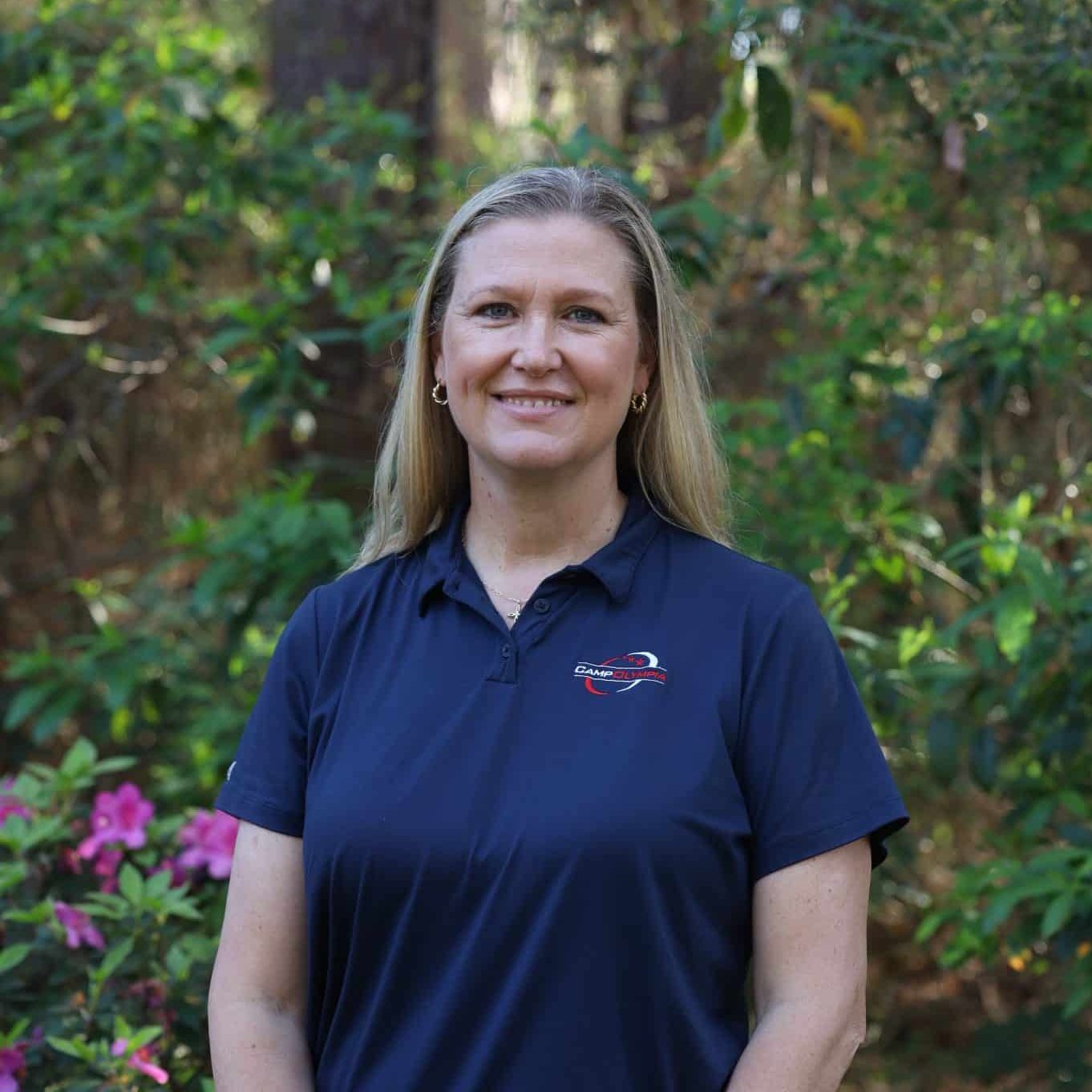 A woman in a navy polo shirt stands smiling in a garden with green foliage and pink flowers.