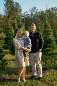 A smiling family of three stands together outdoors among evergreen trees on a sunny day.